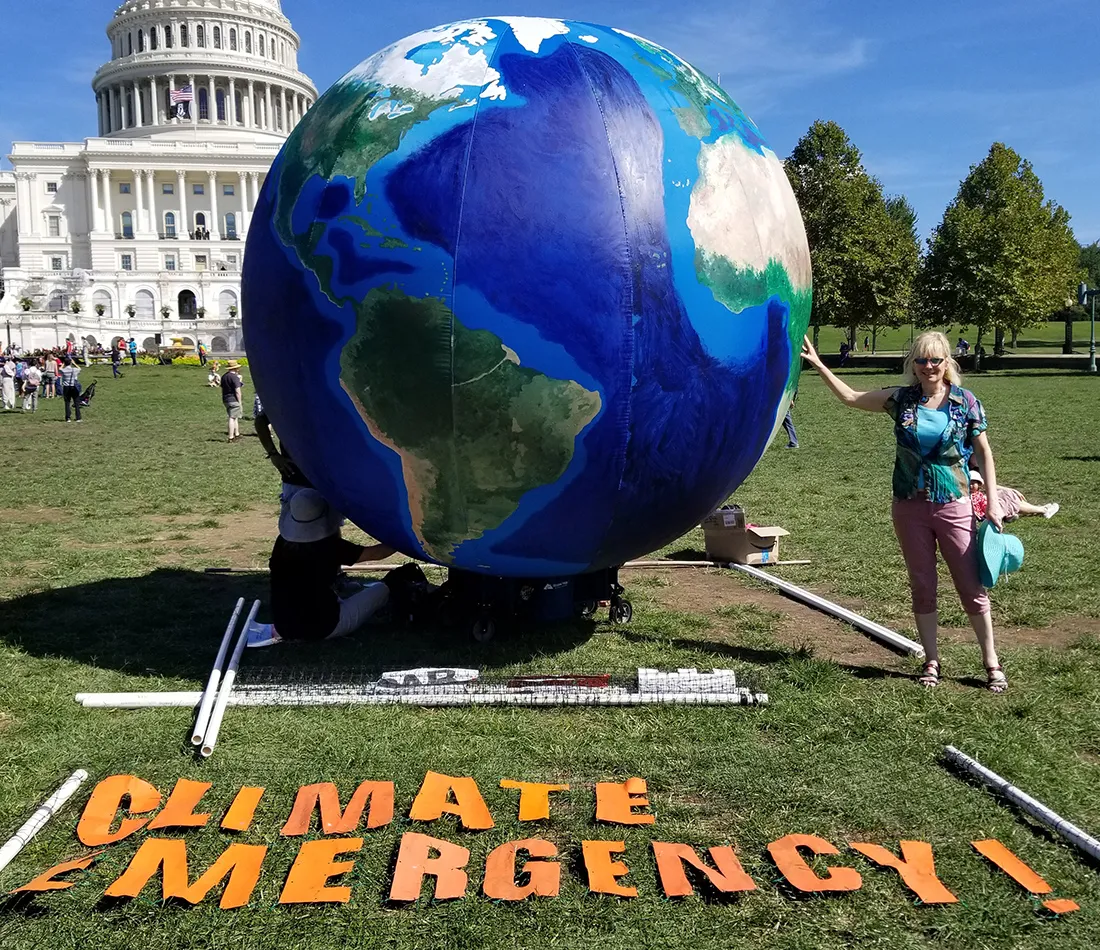 A woman standing with one hand touching a huge Earth beach ball in front of the U.S. Capitol building. Cut-out letters in front of the ball spell "Climate Emergency."