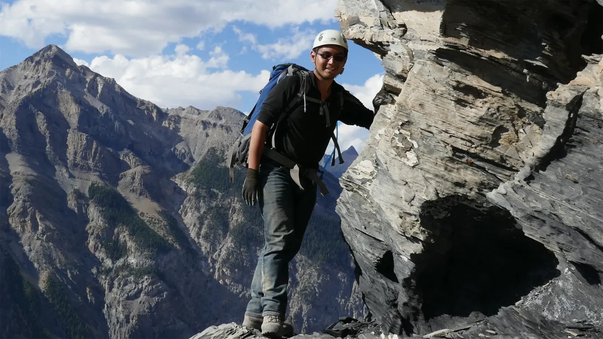 A man leaning against a rock face while wearing a helmet and backpack. Mountains are in the background.
