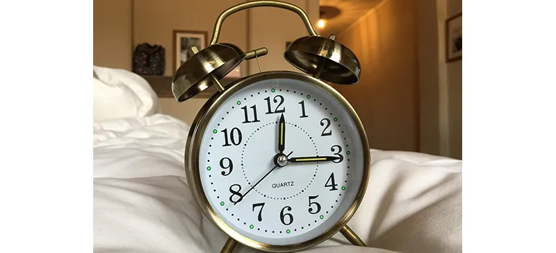 Round, brass analog clock with two brass alarm bells on top. The clock hands read 12:15.