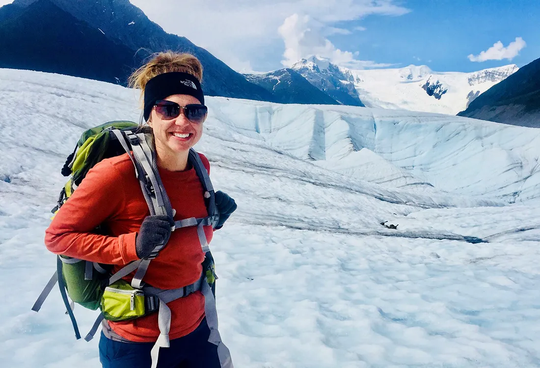 A woman in a red shirt, wearing a backpack and standing in a snowfield with mountains in the distance.