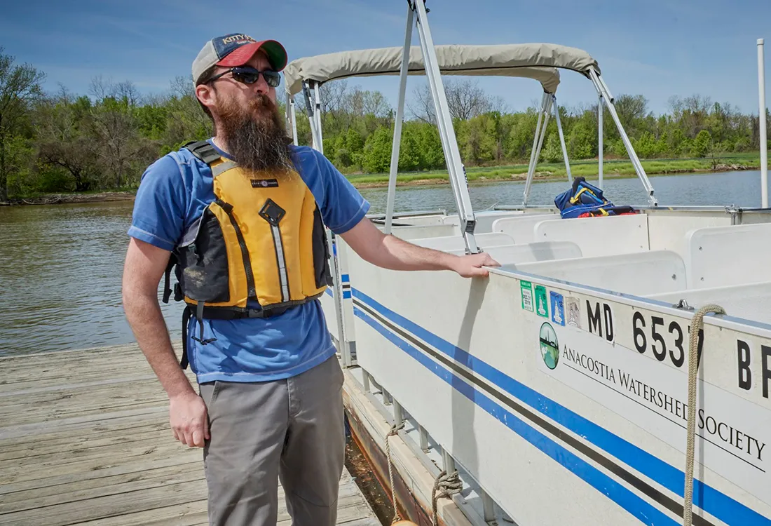 A bearded man wearing a baseball cap and flotation vest stands on a dock next to a small boat.