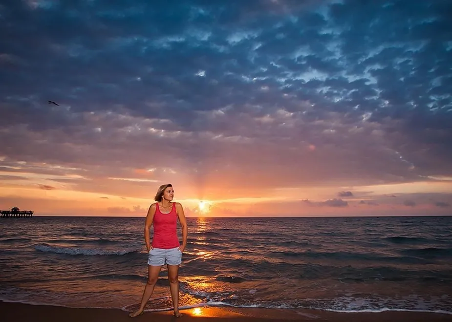 woman in red shirt and white shorts stands in ankle-deep surf on a beach at sunset