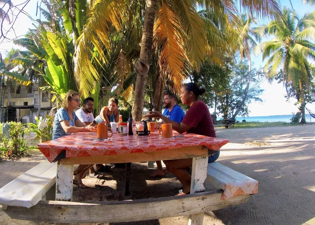 group of people sitting at a picnic table under palm trees on a beach