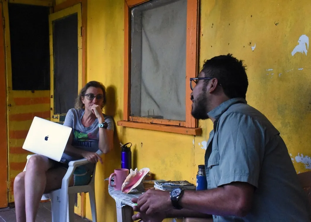 a blonde woman with a laptop listens to a dark-haired man speak on the porch of a yellow building with an orange window frame in the background between them