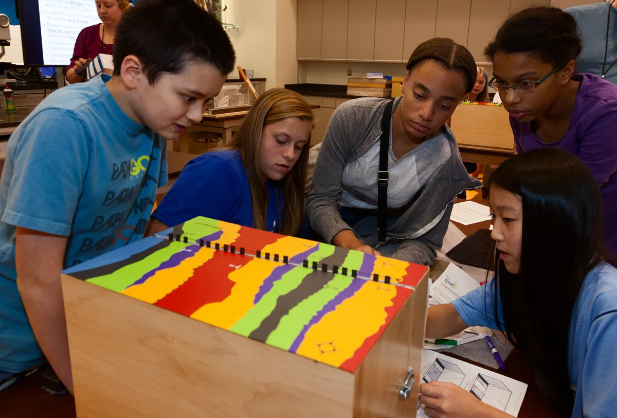 Five teenage students looking intently and interacting with a wooden box with colored stripes on its top