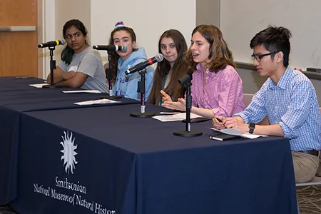 Five teenage students sitting at tables with microphones in front of them. One girl is talking into a microphone.