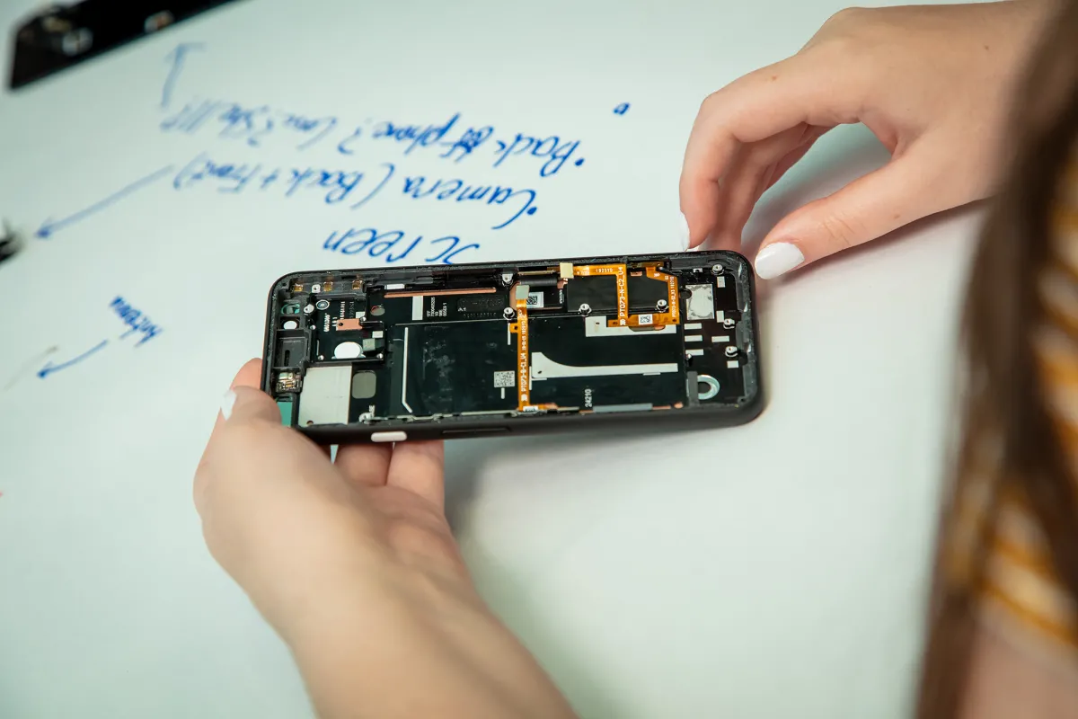 Two hands holding a cellphone that has half of its case removed, exposing the inside. The cellphone is leaning on a table covered with paper that has writing on it about the parts of the cellphone.