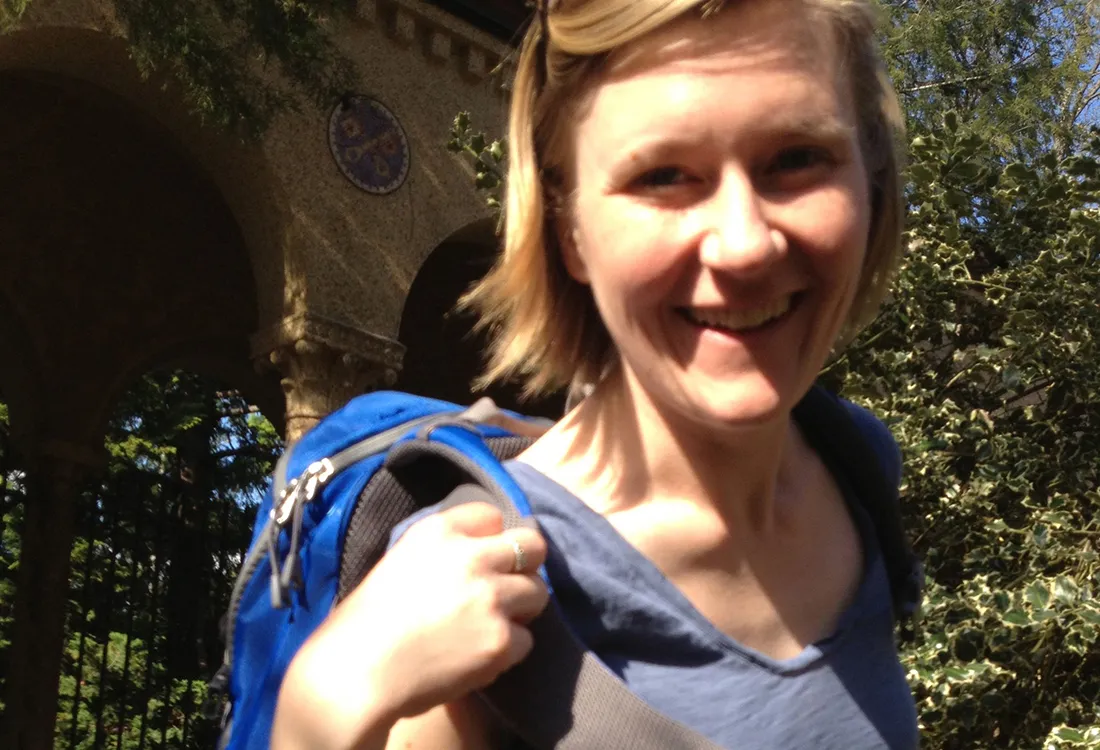 A woman holding a backpack on her shoulder, standing in front of a tree.