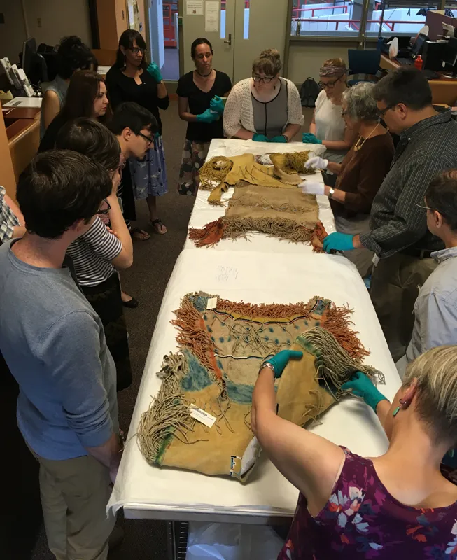 Students gathered around a table, examining 3 Plains Indian shirts