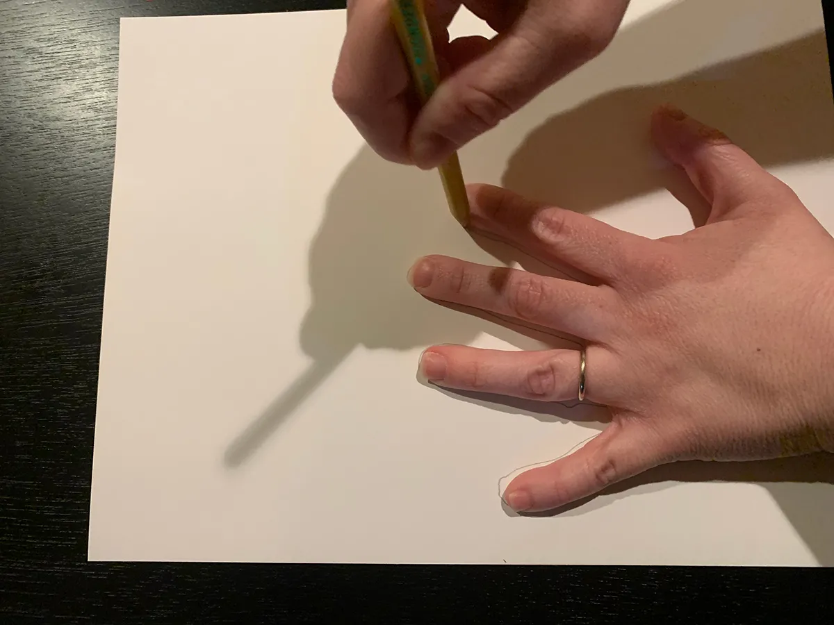 One hand holding a pencil traces another hand that is flat on a piece of paper.