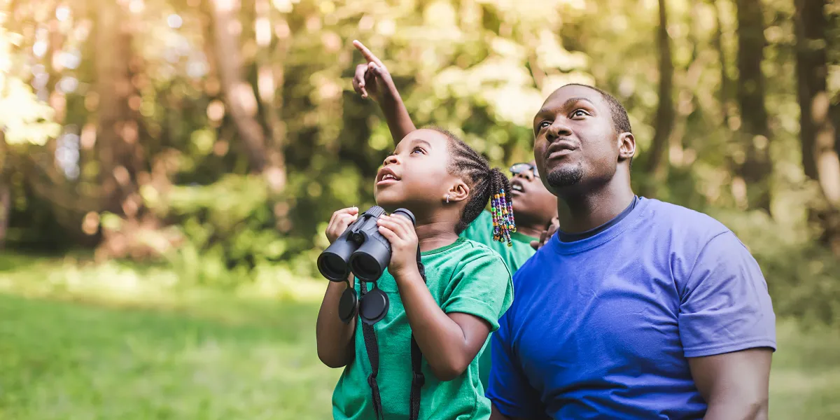 A dark-skinned man and his young daughter and son in front of some trees, looking up as if birdwatching. The girl holds binoculars and the boy is pointing diagonally up.